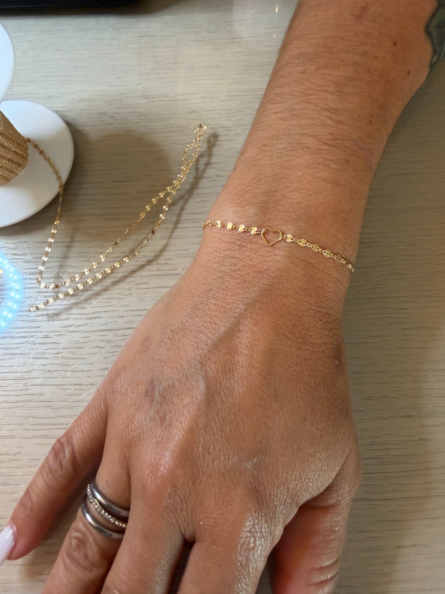 Client's hand with a freshly welded gold heart-link bracelet, chain samples on the table — mobile permanent jewelry by The Bronze Lily, St. Petersburg, FL