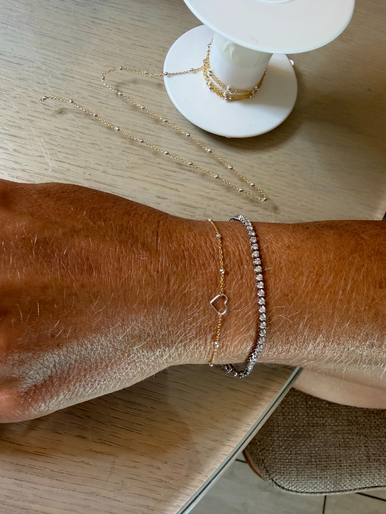 Stacked welded bracelets on a wrist — a tennis-style chain and a dainty gold heart chain — mobile permanent jewelry from The Bronze Lily, St. Petersburg, FL